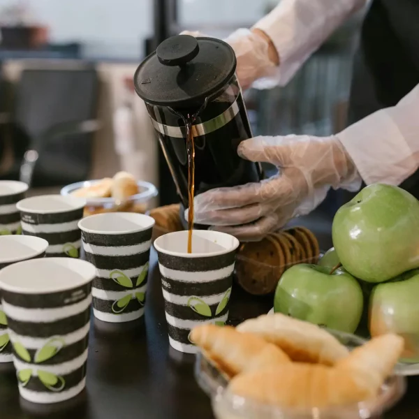 Barista pours coffee from French press into cups at a breakfast event with croissants and apples.